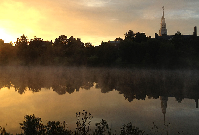 Sunrise over a misty lake, reflecting buildings and trees against a golden sky.