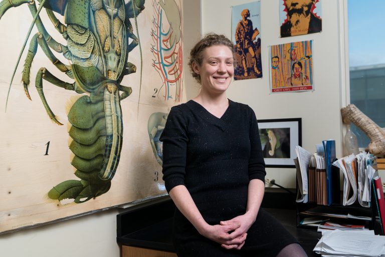 Smiling woman in a black dress stands by a desk with artwork and a large anatomical illustration on the wall behind her.