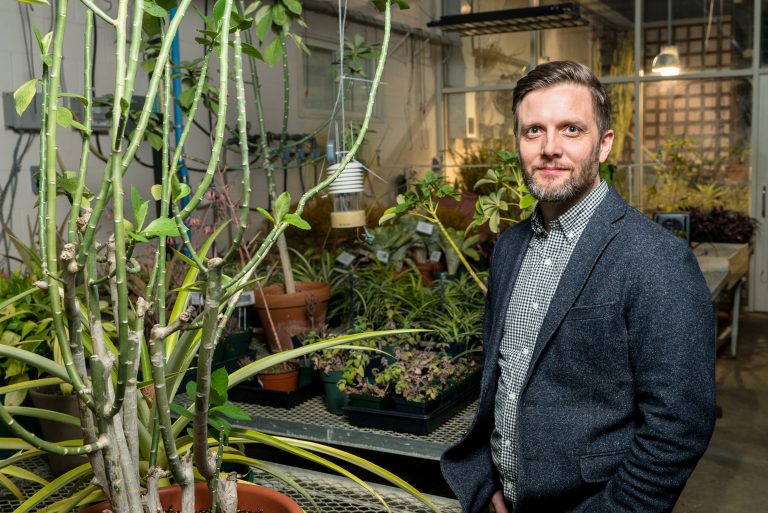 Man in a blazer stands among various plants in a greenhouse, with green foliage and pots visible in the background.