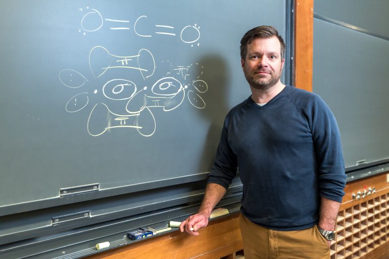 Man standing next to a chalkboard with molecular diagrams and equations drawn on it. Classroom setting.