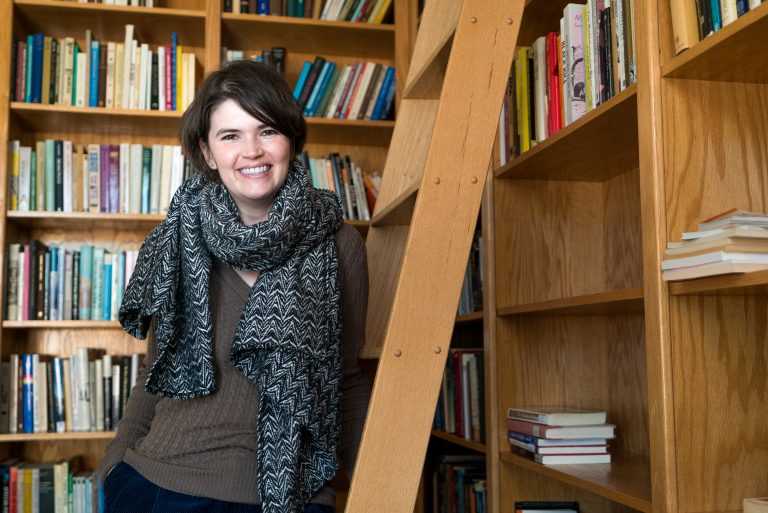 Young woman with short dark hair, wearing a scarf, smiles against a backdrop of wooden bookshelves filled with books.