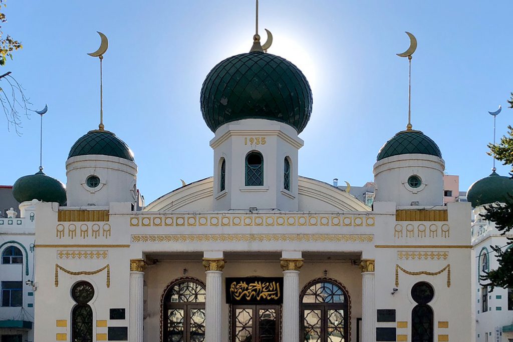 Historic building with green domes and crescents, featuring ornate architecture and sunlight glowing behind it.