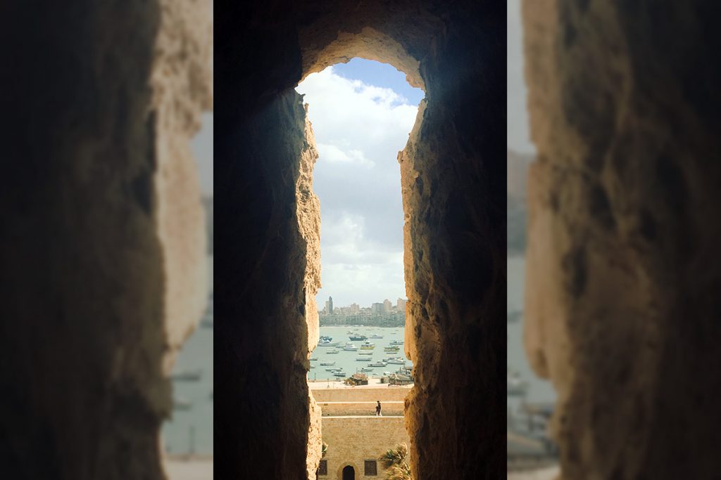 View of a harbor and city skyline through a stone archway, with boats visible on the water under a cloudy sky.
