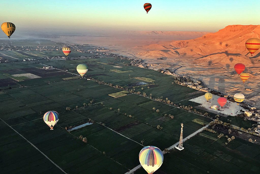 Aerial view of colorful hot air balloons flying over lush green fields and distant hills at sunrise.