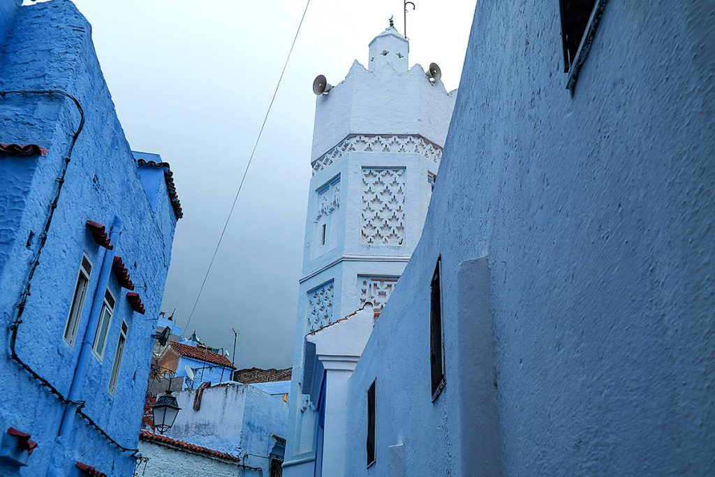 Narrow alleyway with blue-painted buildings leading to a tall, white tower under a cloudy sky.