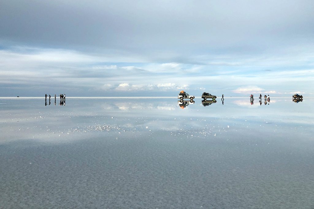 A vast salt flat with reflections of vehicles and people, under a cloudy sky, creating a serene, mirror-like landscape.