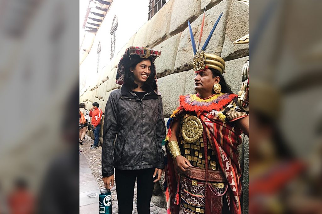 A woman and a man in traditional attire stand in a narrow street, with stone walls and people in the background.
