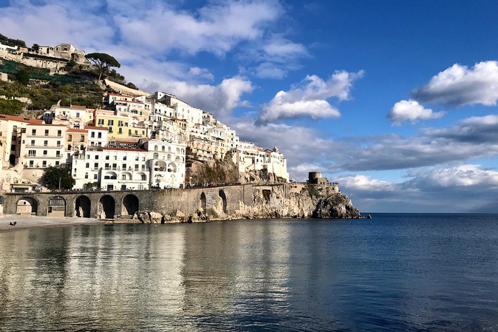 Coastal village with colorful buildings on a hillside, reflecting in calm water under a blue sky with scattered clouds.