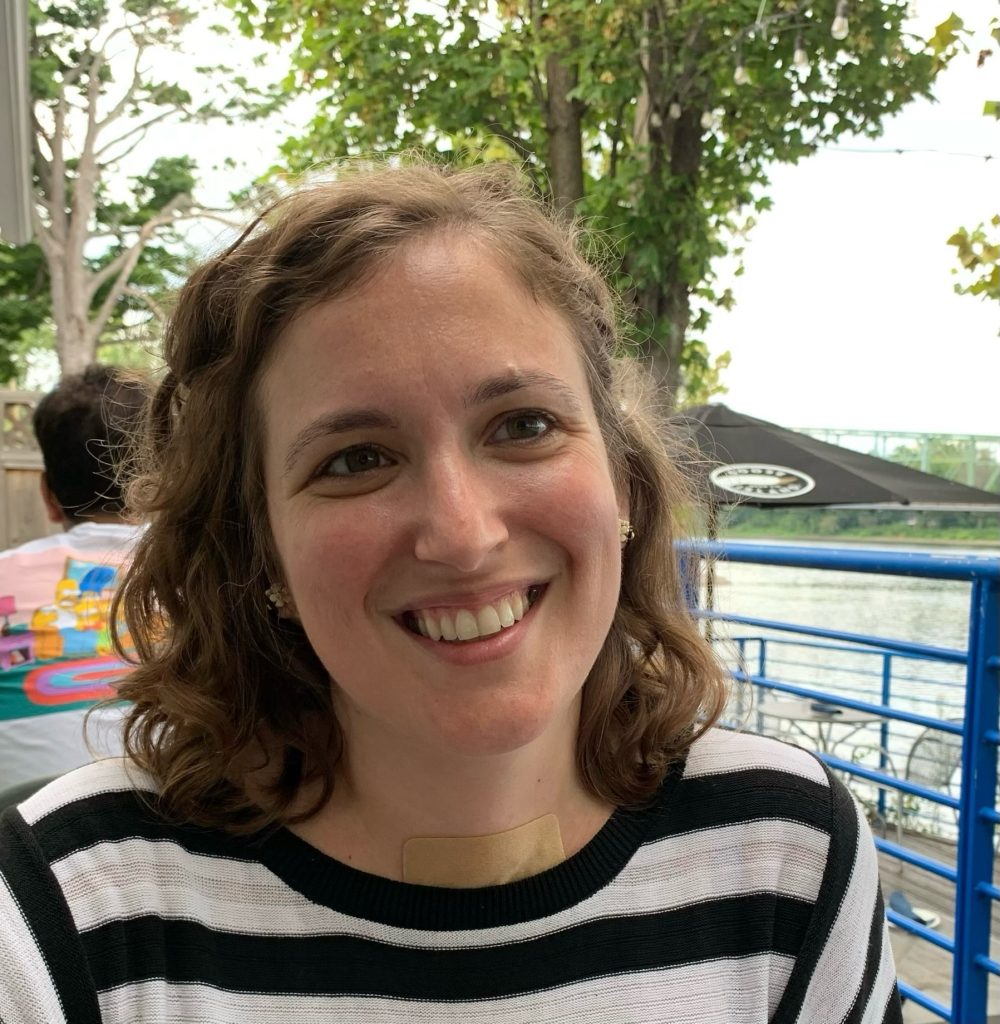 Smiling woman with curly hair wearing a striped shirt, sitting outdoors near a river with greenery in the background.