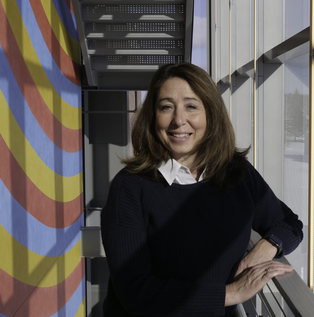 A woman stands by a brightly colored wall, smiling, with natural light illuminating the modern interior space around her.