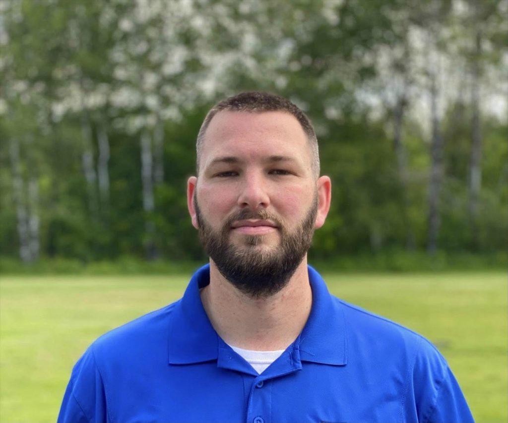 A man with a beard wearing a blue polo shirt stands outdoors against a green background of trees.