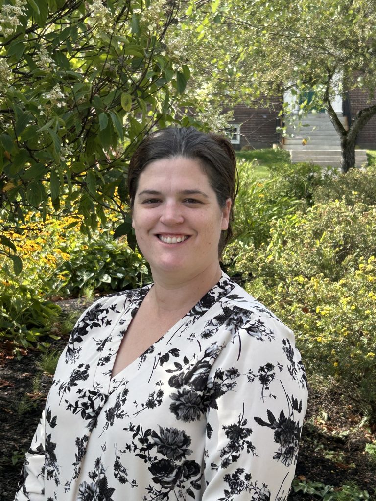 A woman in a floral blouse stands outdoors in a garden with greenery and flowers in the background, smiling at the camera.