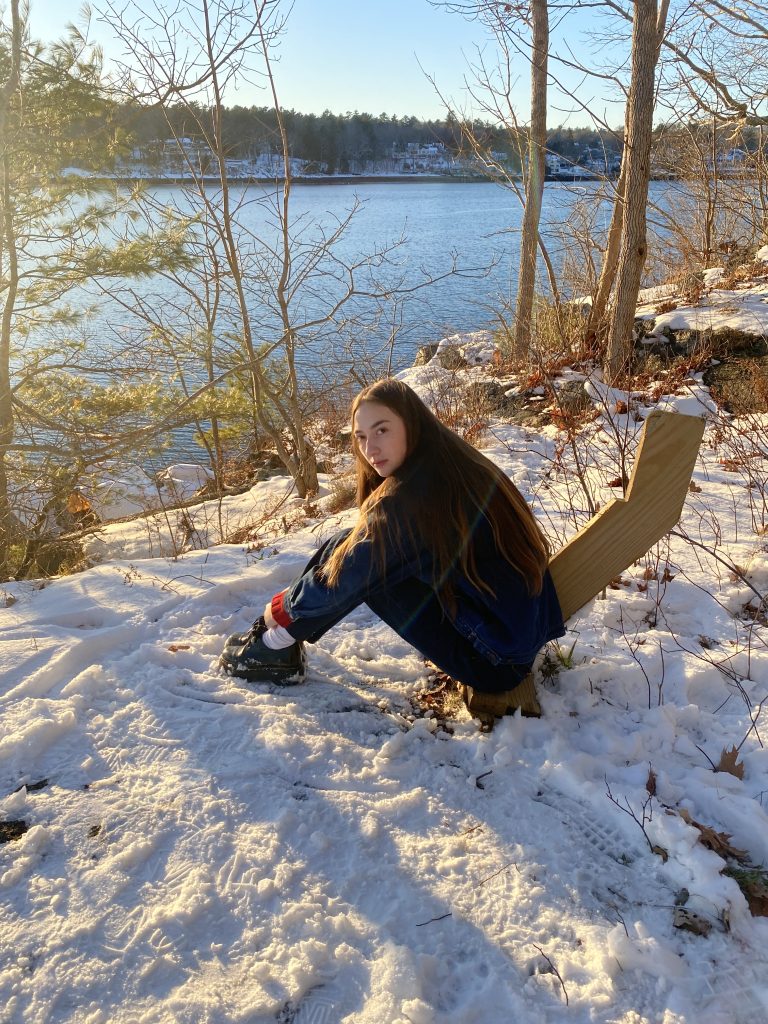 A young woman sits on a snowy riverbank, looking back at the camera with trees and water in the background, capturing a serene moment.