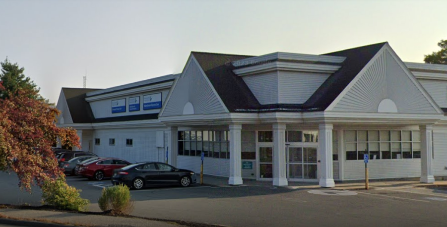 Front view of a large commercial building with a peaked roof and multiple windows, surrounded by parked cars and greenery.