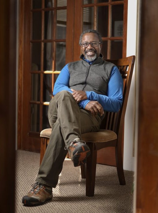 Smiling man sitting on a chair, wearing a blue shirt and gray vest, with brown pants and boots, in a cozy indoor setting.