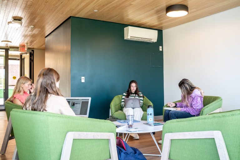 Four people casually working on laptops in a modern, bright lounge area with green chairs and wooden accents.