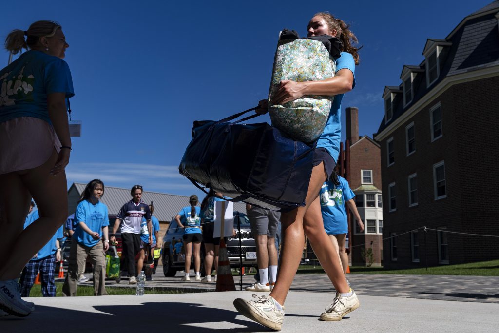 A student carries bags while moving into a college dorm, with others walking nearby on a sunny day.
