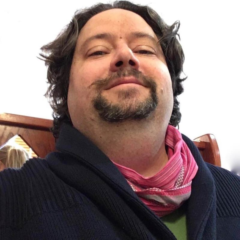A man with curly hair and a goatee, wearing a dark sweater and a pink scarf, poses for a photo.