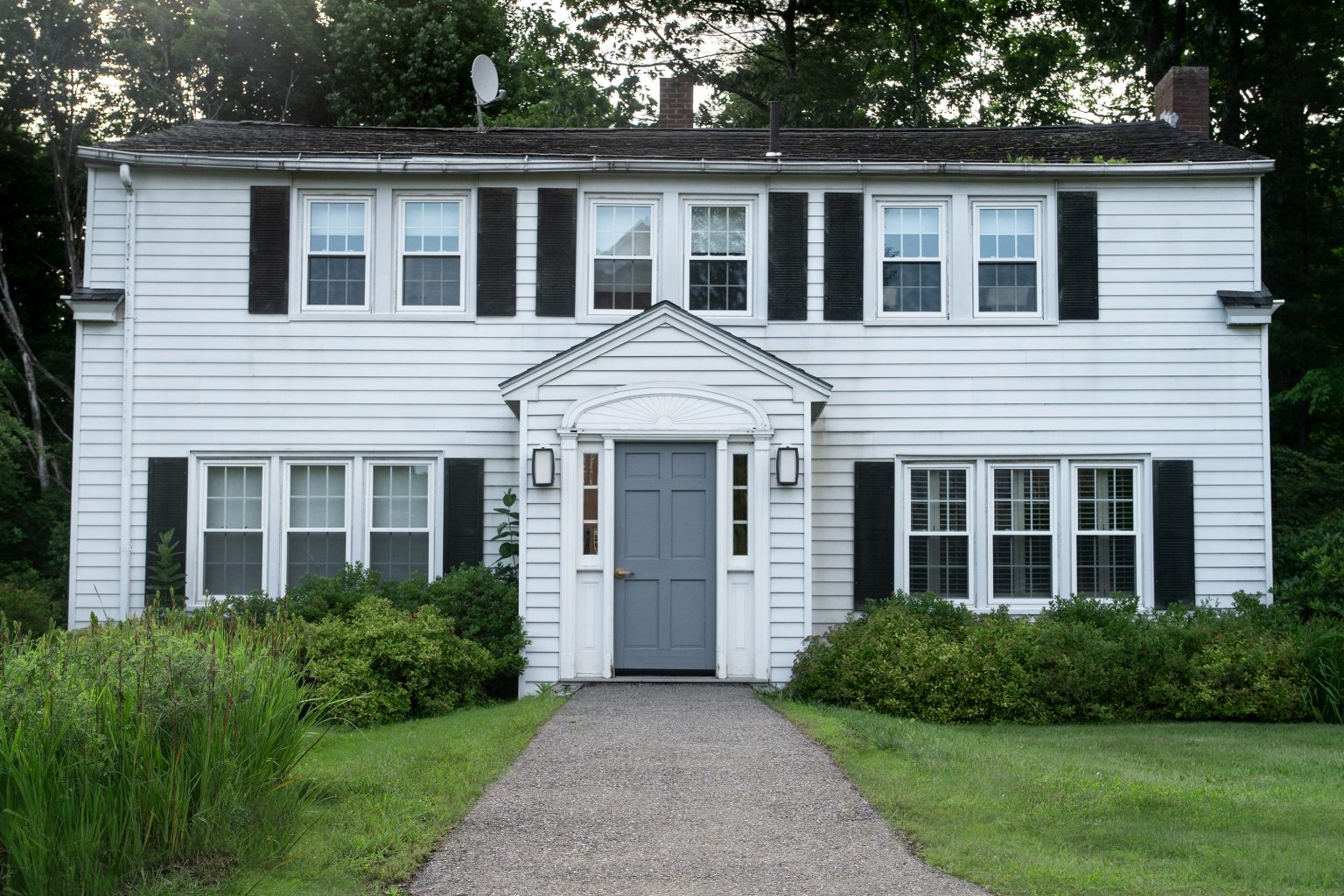 A white two-story house with black shutters, a gray door, and a landscaped front pathway surrounded by greenery.