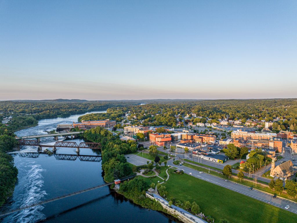 Aerial view of a scenic riverside town with a bridge, waterfront, and green spaces under a clear sky at sunset.