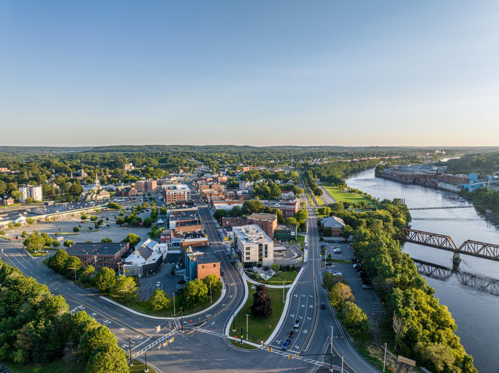 Aerial view of a river city with green hills, roads, buildings, and a visible bridge crossing the river.