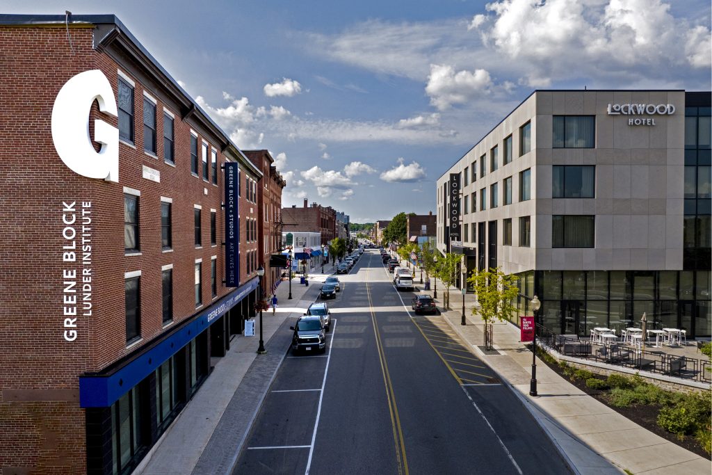 View of a lively street featuring Greene Block Under Institute and Lockwood Hotel, with clear skies and parked cars.