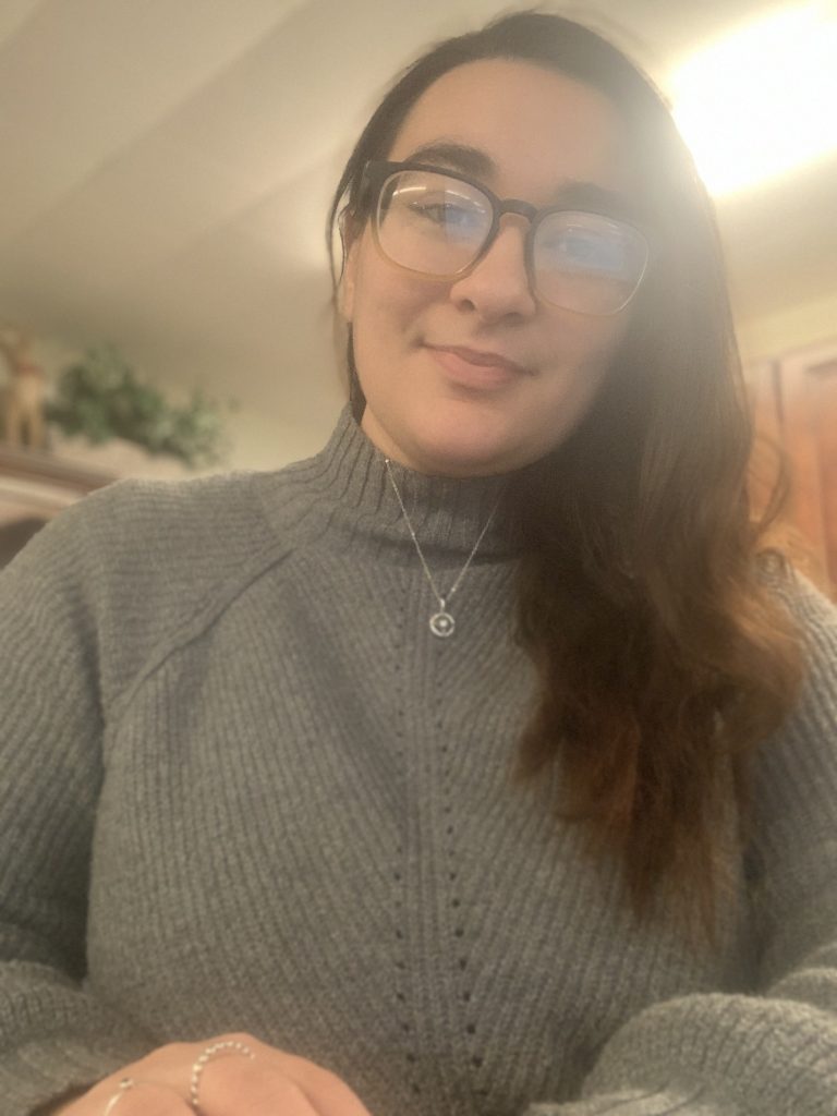 A smiling person with long dark hair, wearing glasses and a gray sweater, sitting at a desk in a cozy indoor setting.