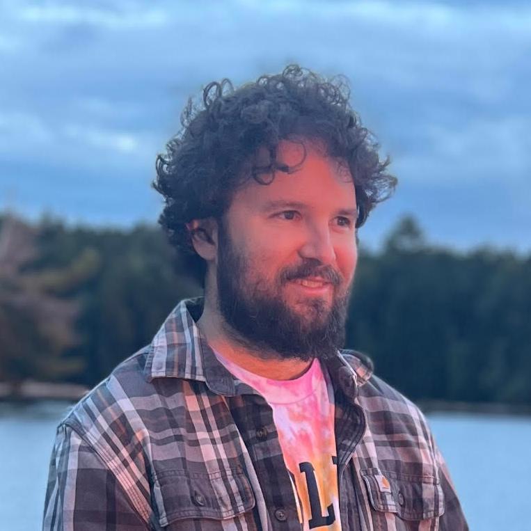 A person with curly hair and a beard smiles against a scenic backdrop of water and trees during sunset.
