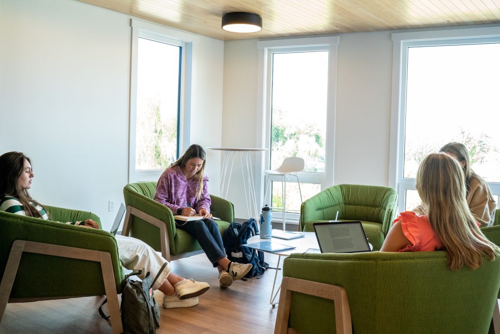 Four people are sitting in green chairs in a bright room with large windows, engaged in study or discussion.