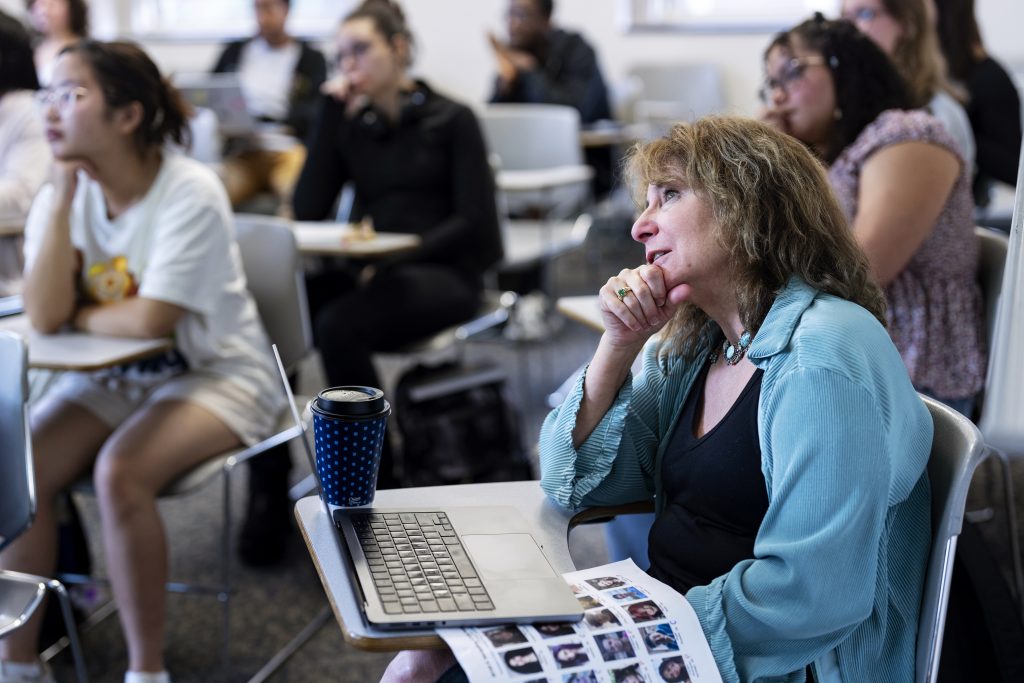A woman sits in a classroom, thoughtful, with a laptop and coffee cup, surrounded by attentive students.