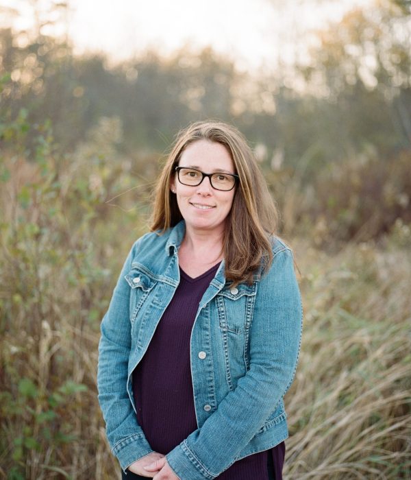 Woman in a denim jacket standing outdoors among tall grass, smiling softly, with a natural background of trees and sunlight.
